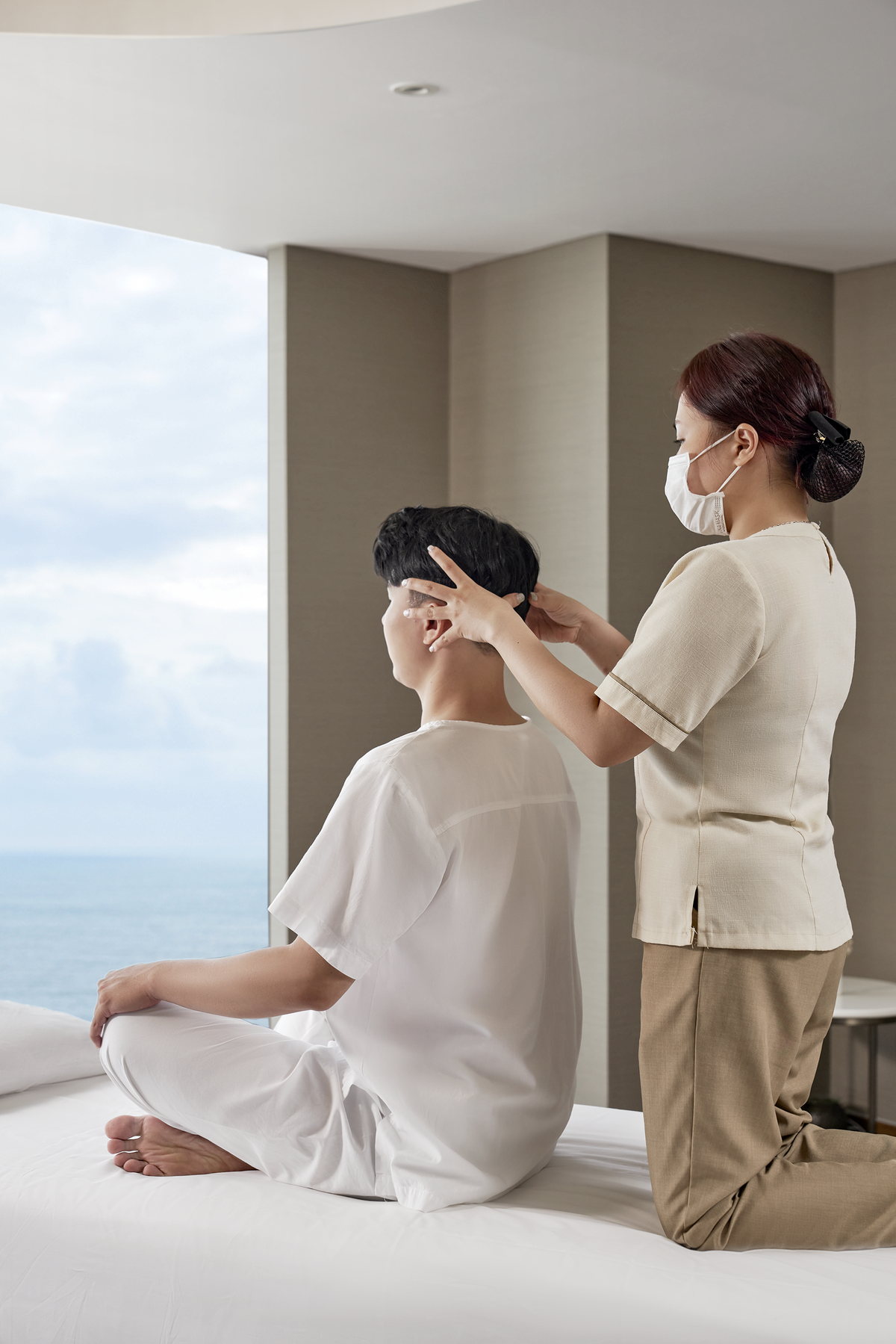 Trị liệu viên massage đầu cho một vị khách ngồi bên cửa kính hướng biển