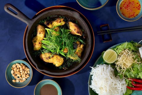 Chảo cá nướng với rau thơm và bún trên nền xanh đậm